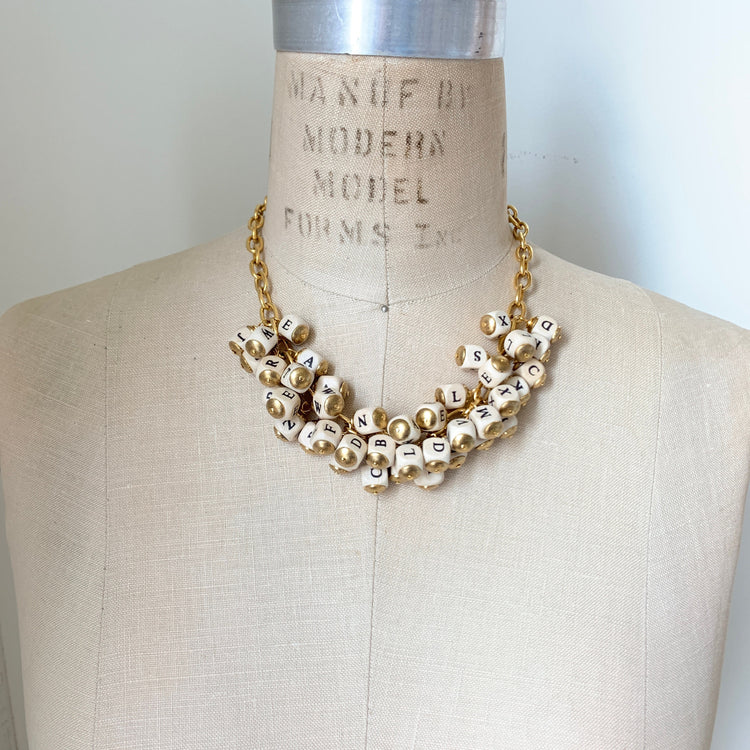 May include: A gold chain necklace with white beads that have black letters on them. The letters spell out the alphabet. The necklace is displayed on a white mannequin. Lenora Dame necklace.