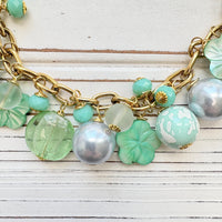 A mix of light green and light blue beads, pearls, and mother of pearl flowers hand from a gold chain necklace shown on a white washed wooden board. Green beaded necklace for women made in the USA. Lenora Dame necklace.