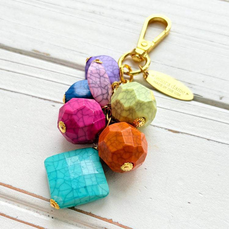 Colorful beaded bag charm with gold tone trigger clasp and brass oval hang tag that says Lenora Dame Made in USA. Bag charm has a mix of different shaped beads in vibrant colors with a black cracked design. Lenora Dame bag charm made in USA.