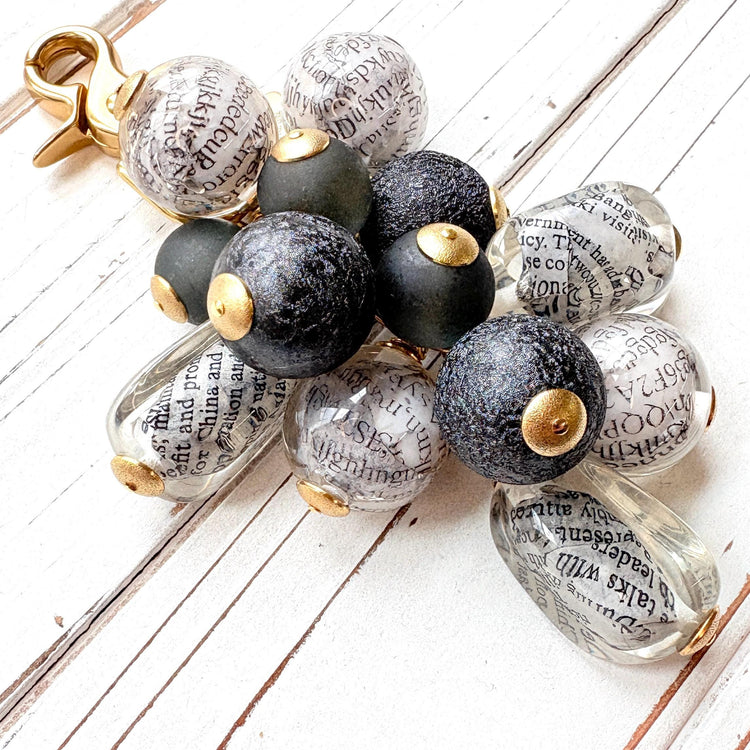 Black and white chunky bag charm with irregular shaped Lucite clear beads with a newspaper print inside hang with round black foil beads and black glass beads in a cluster under a gold trigger clasp. Lenora Dame bag charm.