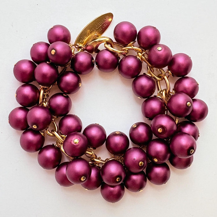 A gold chain bracelet with multiple strands of dark purple faux pearls. The bracelet has a small tag that reads 'Lenora Dame' and 'Made in the USA.'