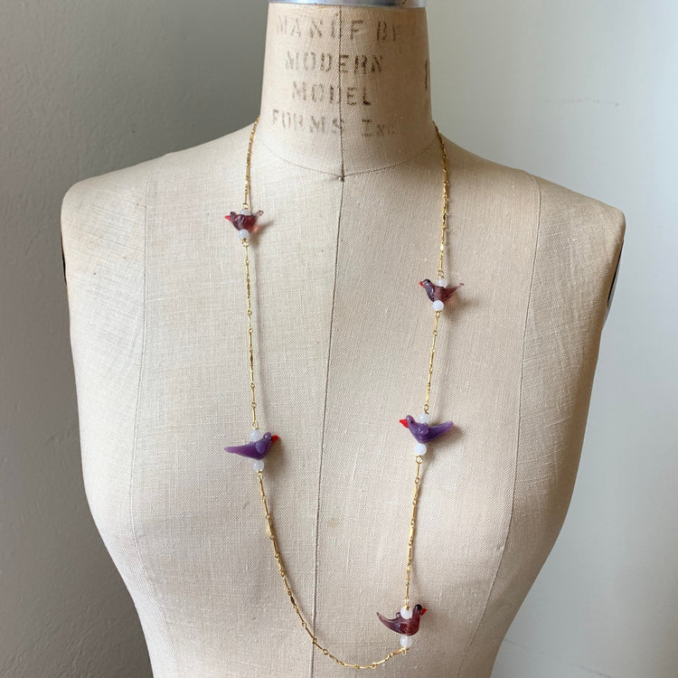 Little purple glass lampworked birds spaced out between bits of gold plated bar chain to create a long necklace. Shown on a vintage dress making form.