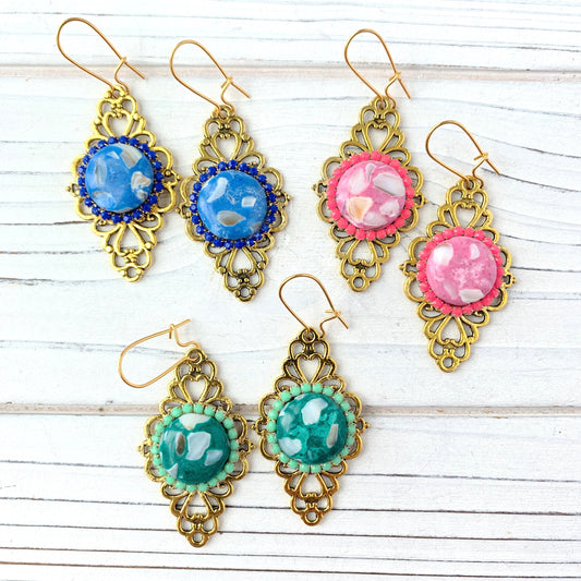 May include: Three pairs of gold filigree earrings with different colored cabochon stones. The earrings are displayed on a white wooden surface. The stones are blue, pink, and green.