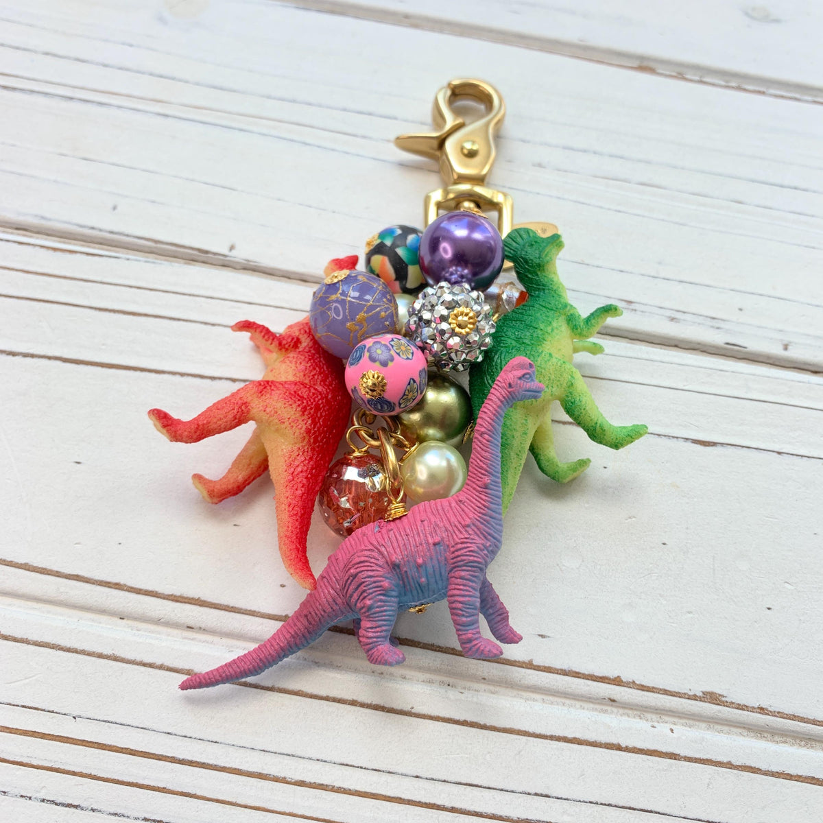 Cute bag charm with red dinosaur, green dinosaur, and pink dinosaur figurines hanging with a mix of colorful beads on a gold chain with a brass trigger clasp. Lenora Dame bag charm.