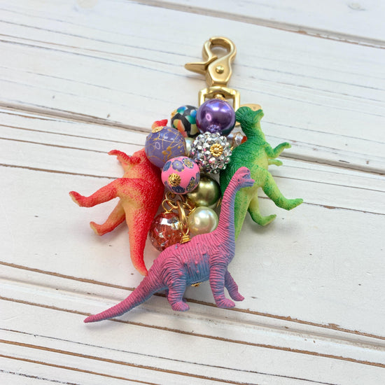 Cute bag charm with red dinosaur, green dinosaur, and pink dinosaur figurines hanging with a mix of colorful beads on a gold chain with a brass trigger clasp. Lenora Dame bag charm.
