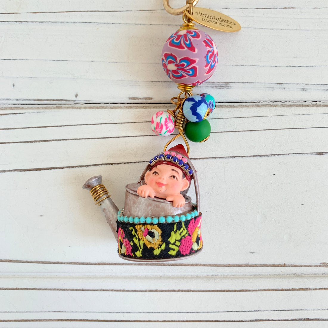 May include: A whimsical charm featuring a small figurine of a child sitting inside a silver watering can with a colorful floral design. The charm is attached to a gold chain with a pink, blue, and green patterned bead and a tag that reads &