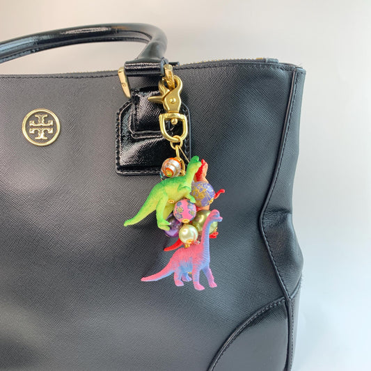 Dinosaur bag charm with a pink dinosaur, red dinosaur, and green dinosaur figurines and a mix of colorful beads and pearls on a gold chain with gold trigger clasp. Lenora Dame bag charm. Cute bag charms for handbags.