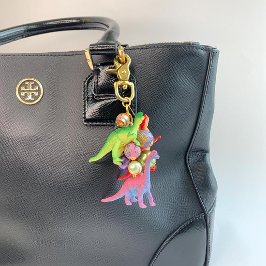 Dinosaur bag charm with a pink dinosaur, red dinosaur, and green dinosaur figurines and a mix of colorful beads and pearls on a gold chain with gold trigger clasp. Lenora Dame bag charm. Cute bag charms for handbags.