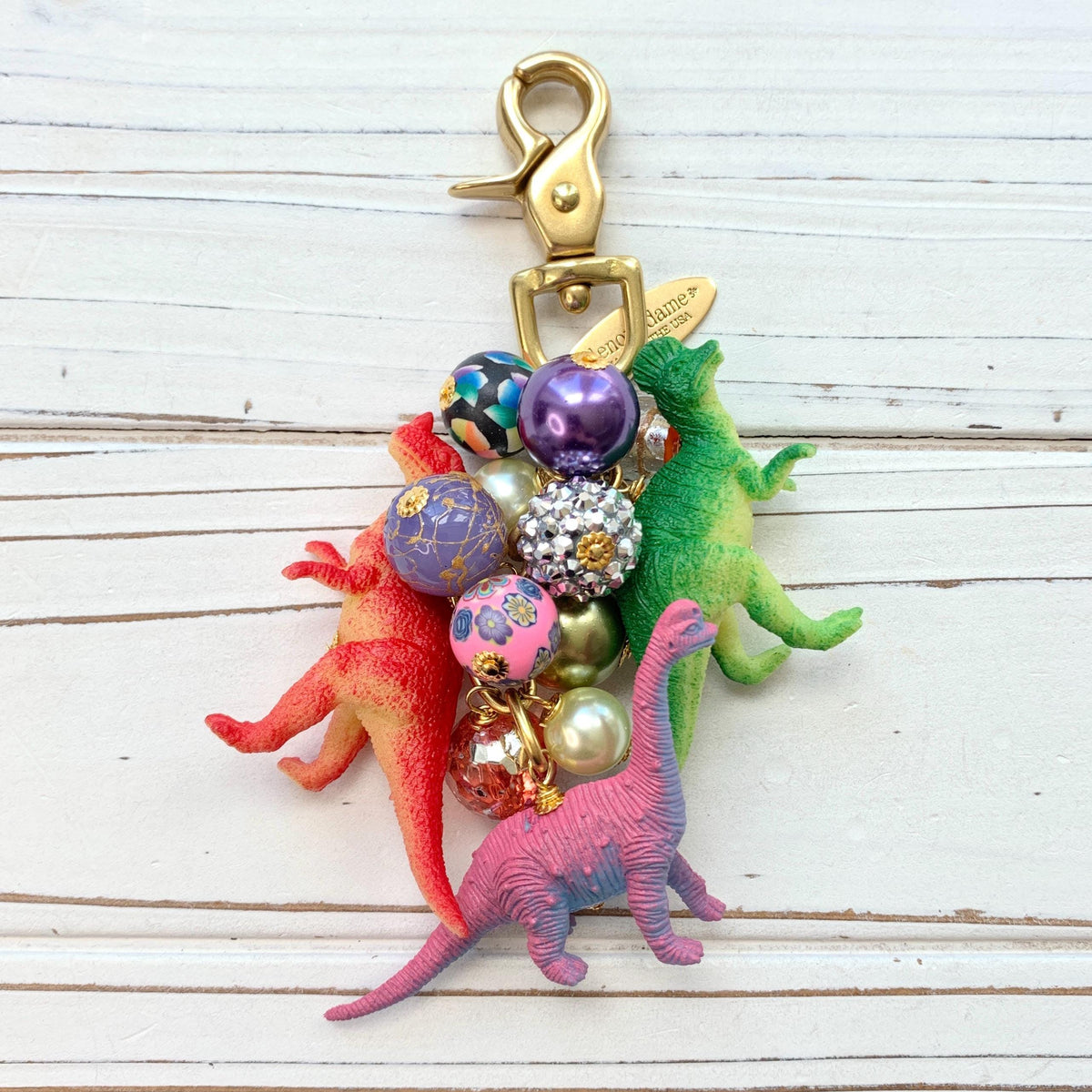 Cute bag charm with red dinosaur, green dinosaur, and pink dinosaur figurines hanging with a mix of colorful beads on a gold chain with a brass trigger clasp. Lenora Dame bag charm.