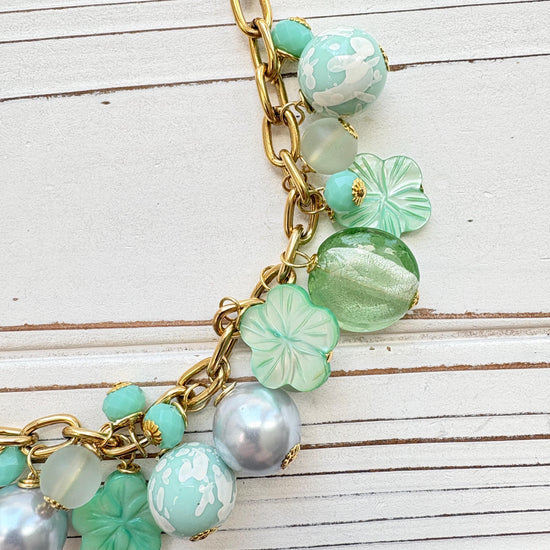 A mix of light green and light blue beads, pearls, and mother of pearl flowers hand from a gold chain necklace shown on a white washed wooden board. Green beaded necklace for women made in the USA. Lenora Dame necklace.