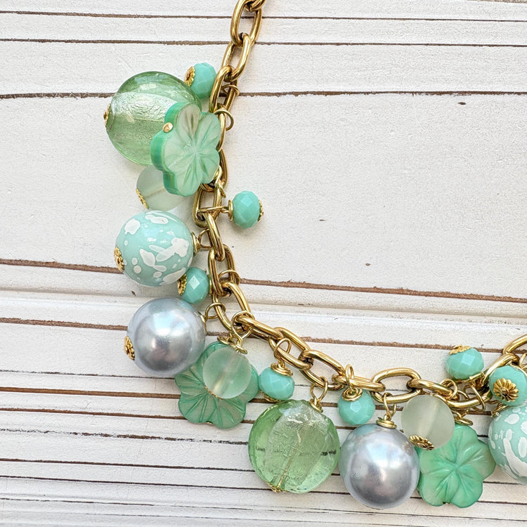 A mix of light green and light blue beads, pearls, and mother of pearl flowers hand from a gold chain necklace shown on a white washed wooden board. Green beaded necklace for women made in the USA. Lenora Dame necklace.