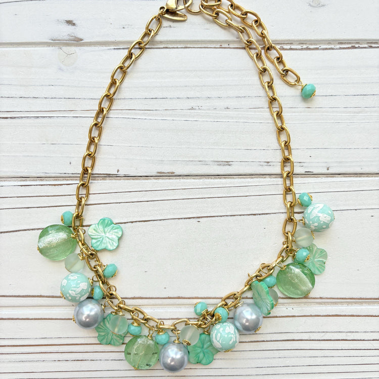 A mix of light green and light blue beads, pearls, and mother of pearl flowers hand from a gold chain necklace shown on a white washed wooden board. Green beaded necklace for women made in the USA. Lenora Dame necklace.