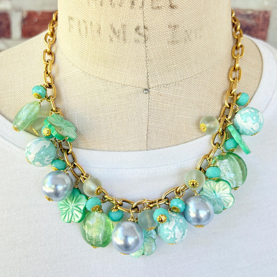A mix of light green and light blue beads, pearls, and mother of pearl flowers hand from a gold chain necklace shown on a dress form wearing a white shirt. Green beaded necklace for women made in the USA. Lenora Dame necklace.