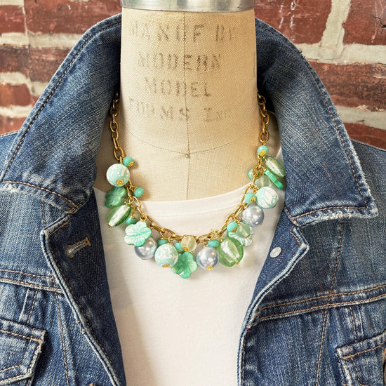 A mix of light green and light blue beads, pearls, and mother of pearl flowers hand from a gold chain necklace shown on dress form wearing a white shirt and dark blue denim jacket with a red brick wall background. Green beaded necklace for women made in the USA. Lenora Dame necklace.