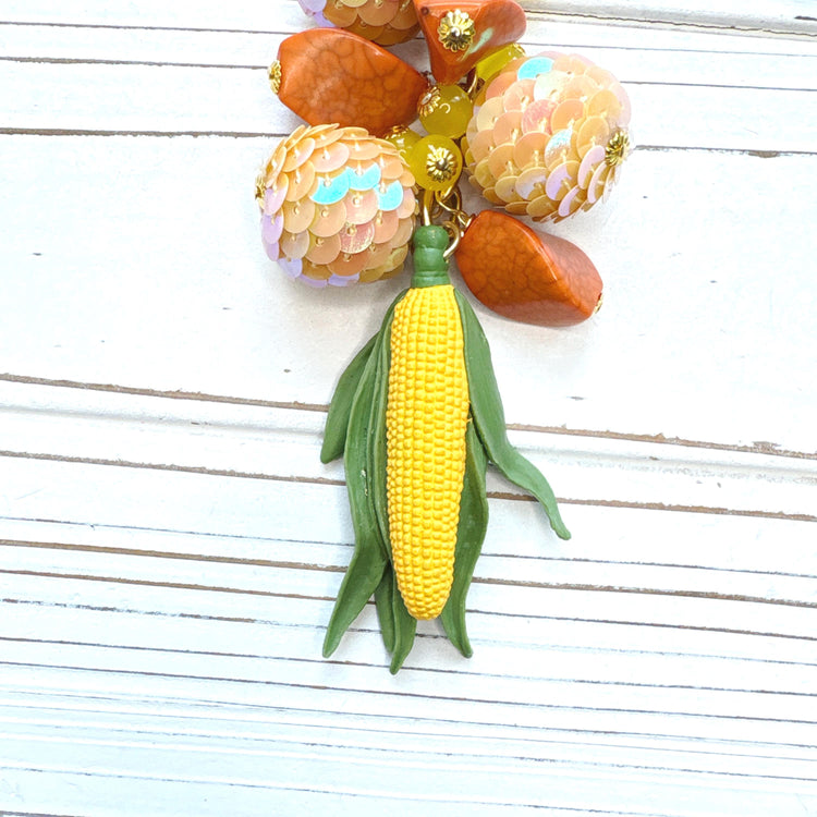 Beaded bag charm with miniature yellow corn cob with green leaves hanging from a mix of different orange beads. Gold trigger clasp at the top with an oval hang tag that says Lenora Dame Made in USA. Lenora Dame bag charm.