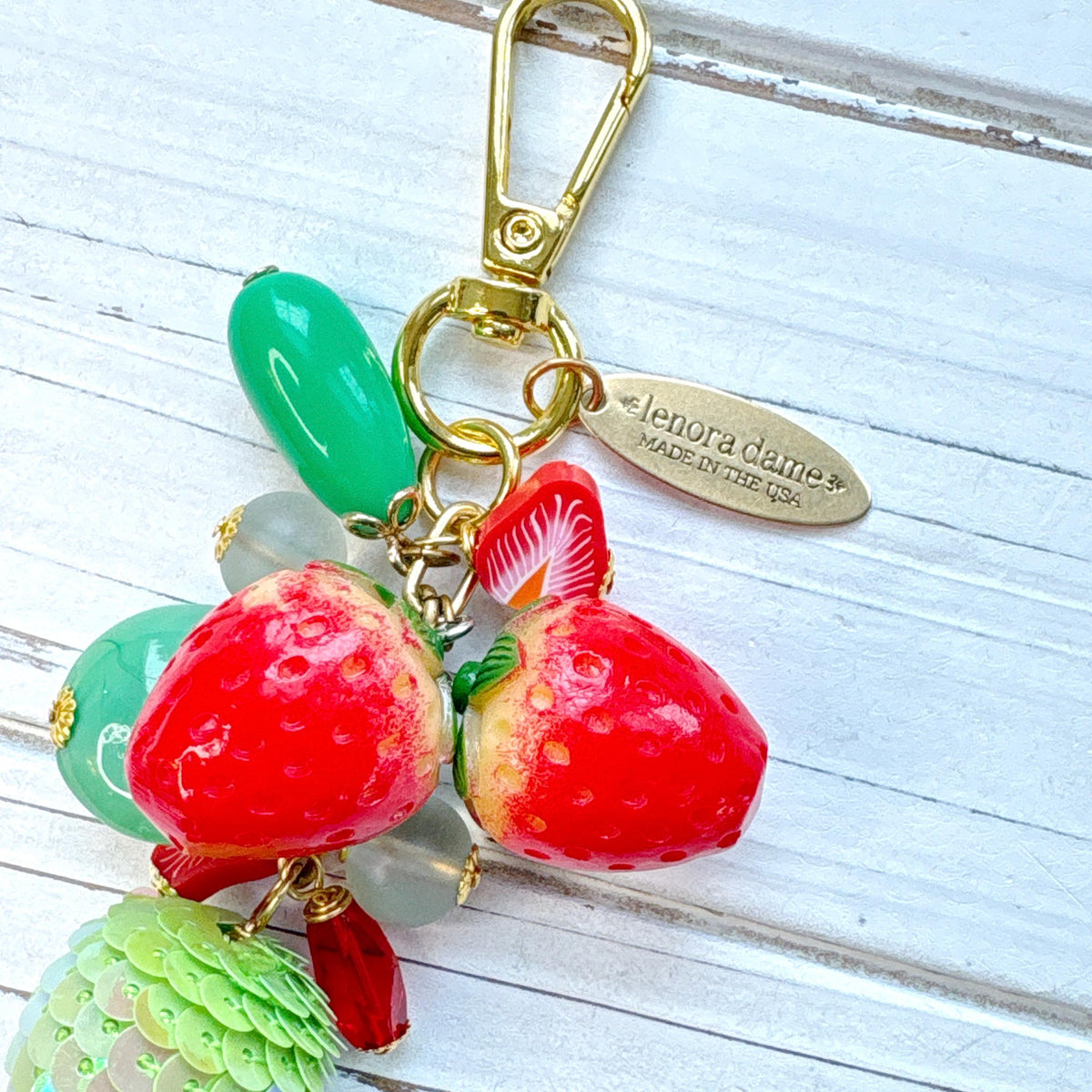 Cute strawberry bag charm with a mix of red and green beads, two red strawberry charms on a gold chain with gold trigger clasp and brass hang tag that says Lenora Dame Made in USA. Lenora Dame bag charm.