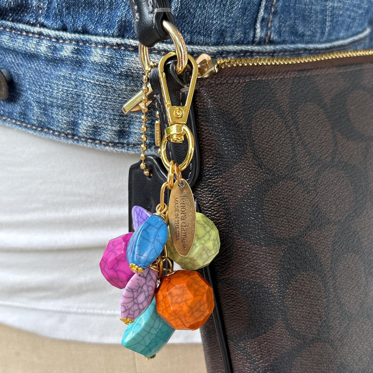Colorful beaded bag charm with gold tone trigger clasp and brass oval hang tag that says Lenora Dame Made in USA. Bag charm has a mix of different shaped beads in vibrant colors with a black cracked design. The bag charm is shown on a black and brown Coach crossbody bag. Lenora Dame bag charm made in USA.