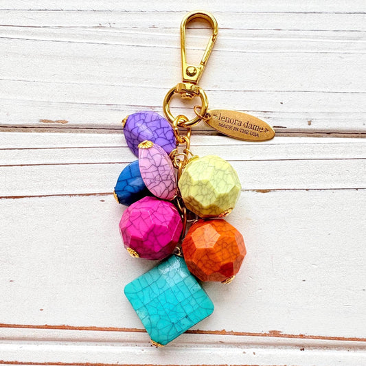 Colorful beaded bag charm with gold tone trigger clasp and brass oval hang tag that says Lenora Dame Made in USA. Bag charm has a mix of different shaped beads in vibrant colors with a black cracked design. Lenora Dame bag charm made in USA.