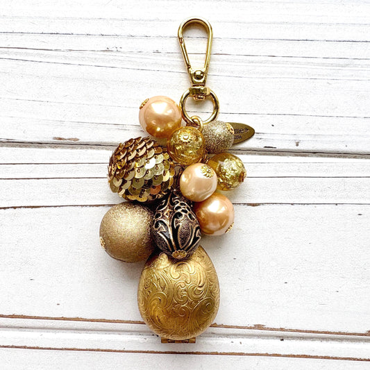 Gold tone beaded bag charm with a etched brass egg shaped hinged photo locket below a mix of pearls, lampworked glass beads, a gold sequin beads, and a black bead with gold scroll design. The beads hang on a gold chain with a gold clasp and Lenora Dame Made in USA hang tag.