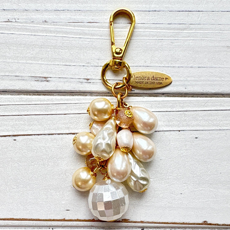 Pearl bag charm shown on a whitewashed board. A mix of cream and white faux pearls in different shapes are wire wrapped to a gold chain with gold clasp and oval hang tag that says Lenora Dame Made in USA. Lenora Dame bag charm.