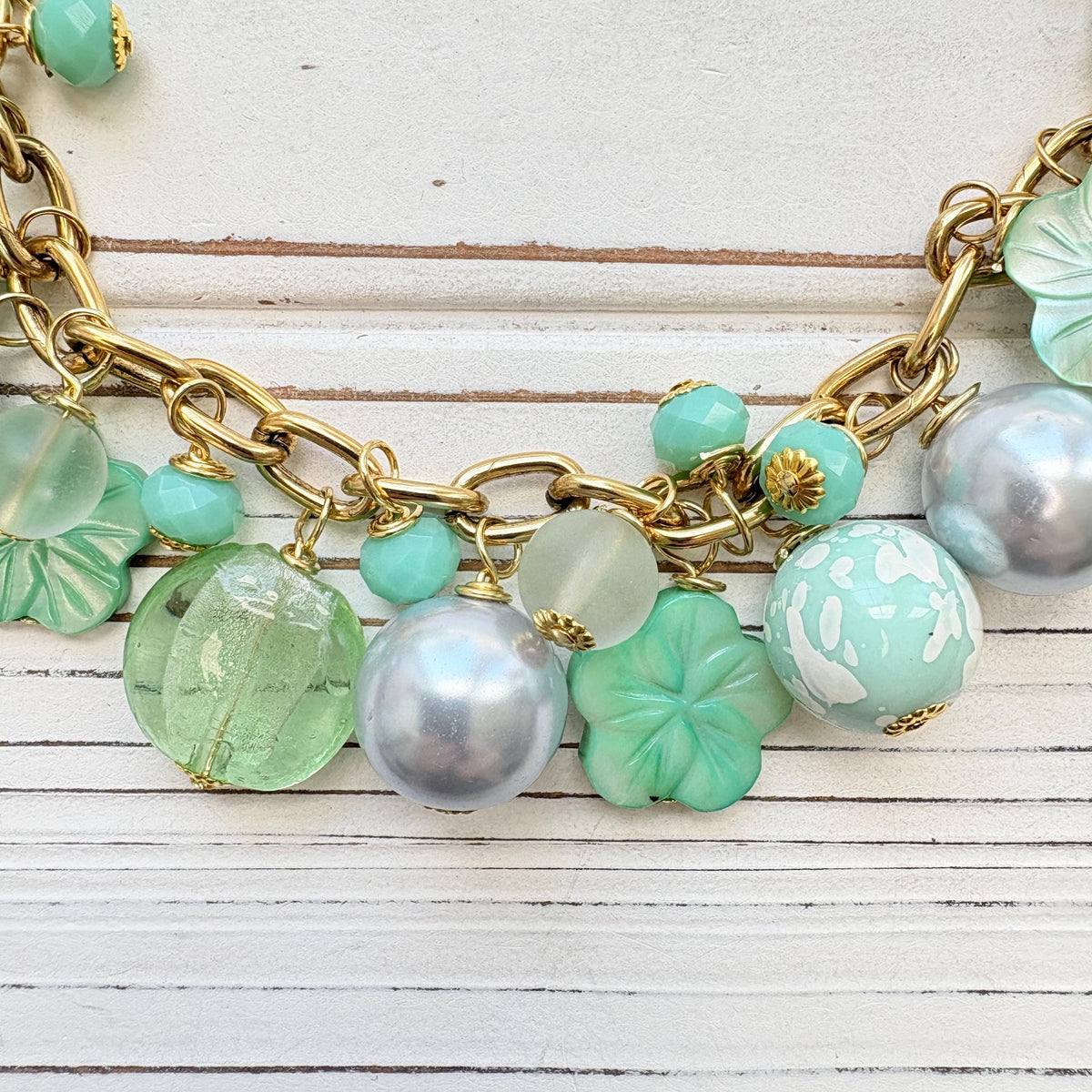 A mix of light green and light blue beads, pearls, and mother of pearl flowers hand from a gold chain necklace shown on a white washed wooden board. Green beaded necklace for women made in the USA. Lenora Dame necklace.