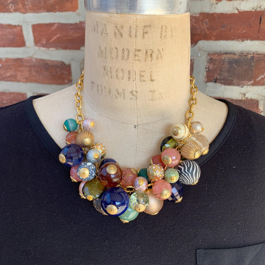 May include: Statement necklace featuring a gold chain and an array of multicolored beads. The beads are varied in size, shape, and pattern, including opaque and translucent textures. Colors include teal, pink, gold, brown, and dark blue. The necklace is displayed on a dress form.