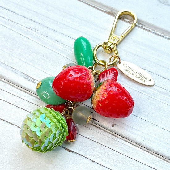 Cute strawberry bag charm with a mix of red and green beads, two red strawberry charms on a gold chain with gold trigger clasp and brass hang tag that says Lenora Dame Made in USA. Lenora Dame bag charm.