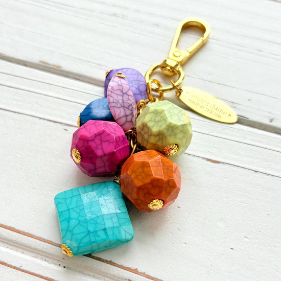 Colorful beaded bag charm with gold tone trigger clasp and brass oval hang tag that says Lenora Dame Made in USA. Bag charm has a mix of different shaped beads in vibrant colors with a black cracked design. Lenora Dame bag charm made in USA.