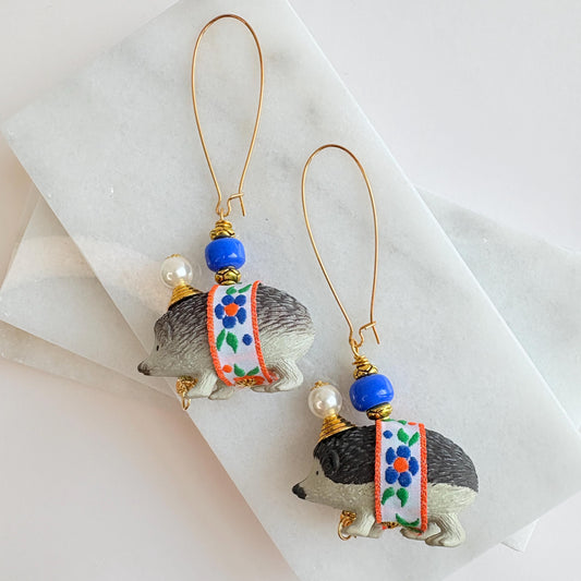 Cute hedgehog earrings with miniature black and gray hedgehog figurines wrapped in blue and orange floral ribbon trim hanging from long gold oval ear wires. Hedgehogs have gold cone and pearl hats and blue beads on their back. Lenora Dame earrings.