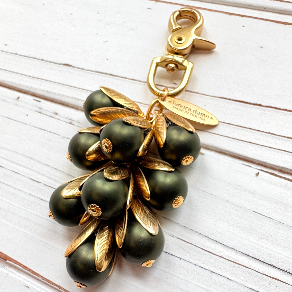 Gold bag charm with a trigger clasp at the top holding a cluster of matte hunter green pearls encased in gold leaf bead caps. A gold oval tag imprinted with Lenora Dame Handmade in USA hangs at the top by the trigger clasp. Lenora Dame bag charms. Green beaded purse charm.
