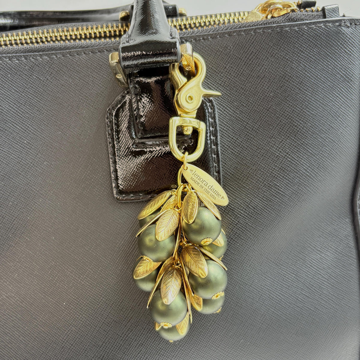 Gold bag charm with a trigger clasp at the top holding a cluster of matte hunter green pearls encased in gold leaf bead caps. A gold oval tag imprinted with Lenora Dame Handmade in USA hangs at the top by the trigger clasp. Lenora Dame bag charms. Green beaded purse charm shown hanging from a black leather handbag strap.