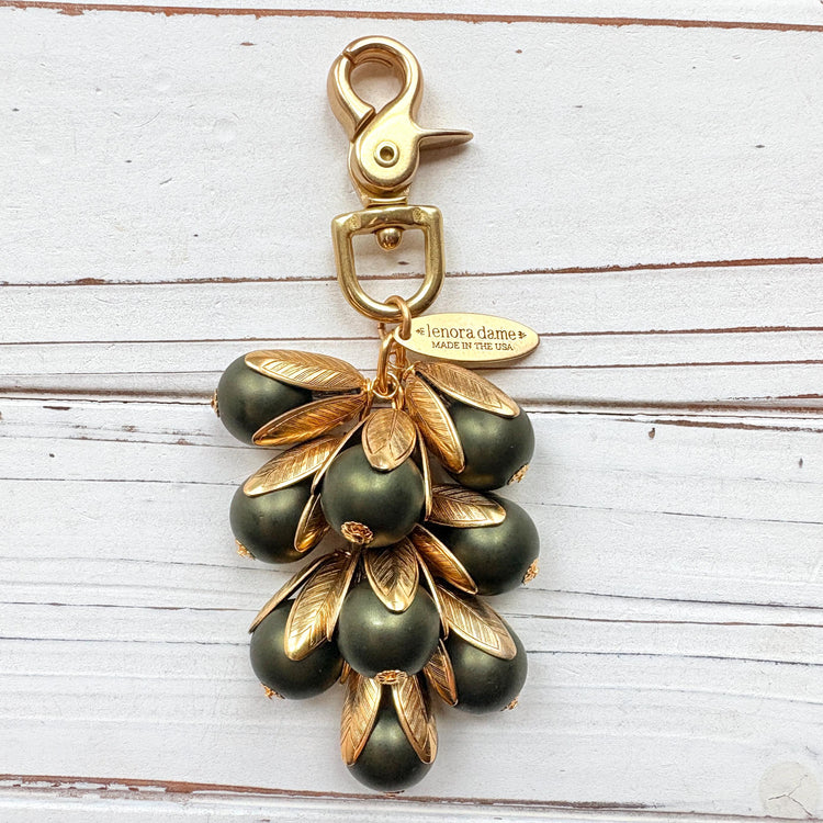 Gold bag charm with a trigger clasp at the top holding a cluster of matte hunter green pearls encased in gold leaf bead caps. A gold oval tag imprinted with Lenora Dame Handmade in USA hangs at the top by the trigger clasp. Lenora Dame bag charms. Green beaded purse charm.