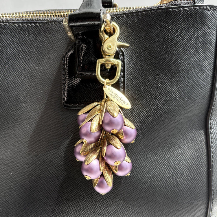 Gold bag charm with a trigger clasp at the top holding a cluster of lavender matte purple pearls encased in gold leaf bead caps. A gold oval tag imprinted with Lenora Dame Handmade in USA hangs at the top by the trigger clasp. Lenora Dame bag charms. Purple purse charm shown hanging on a black leather handbag strap.
