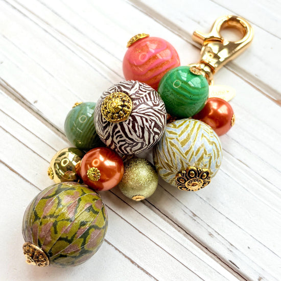 Beaded bag charm with large round beads in a mix of designs in a cluster under a gold trigger clasp. Three beads are wood decoupaged in modern animal prints such as snake print and zebra print. The other beads are gold, rust, green and pink. Lenora Dame bag charm.