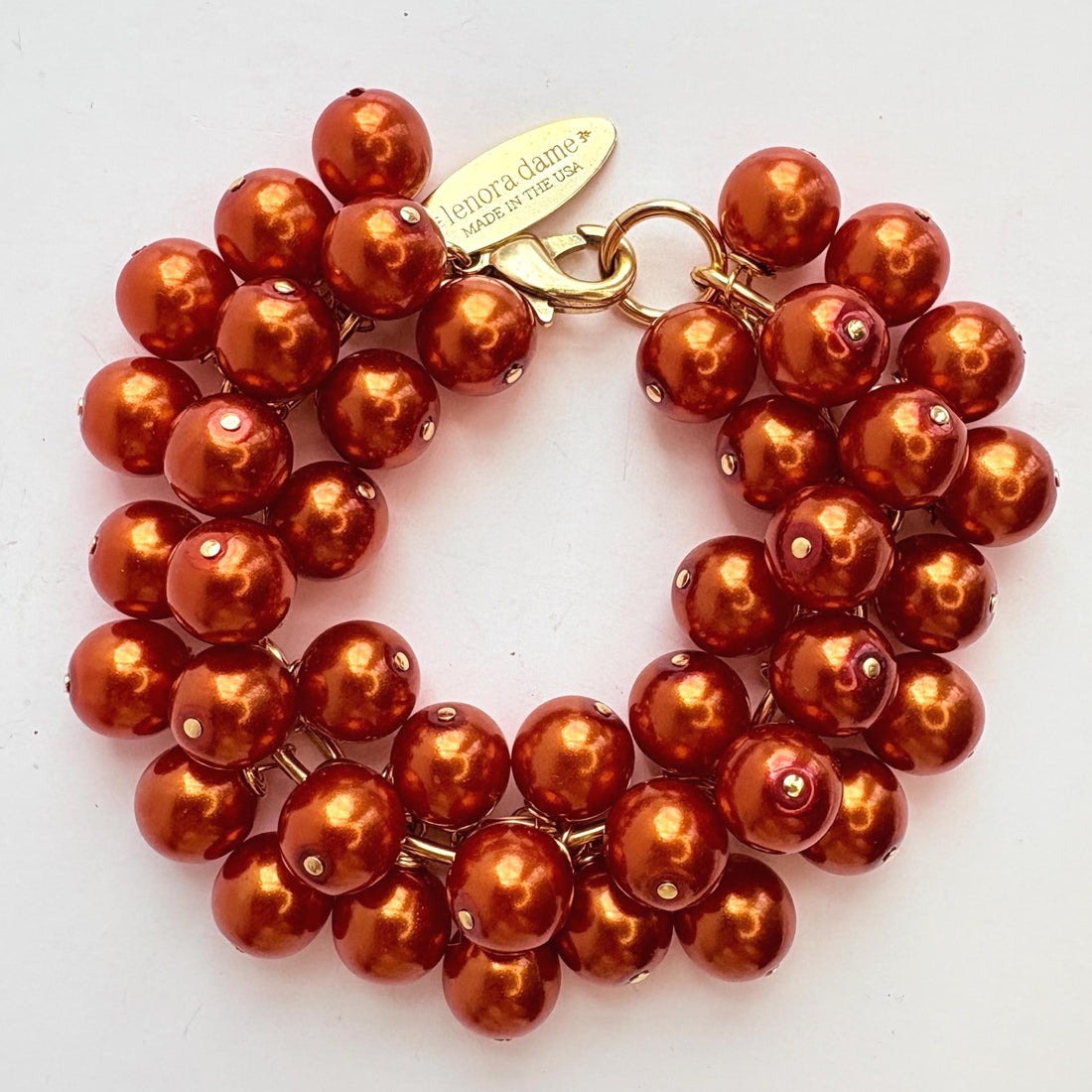 A gold chain bracelet with multiple strands of shiny rust orange faux pearls. The bracelet has a small tag that reads &