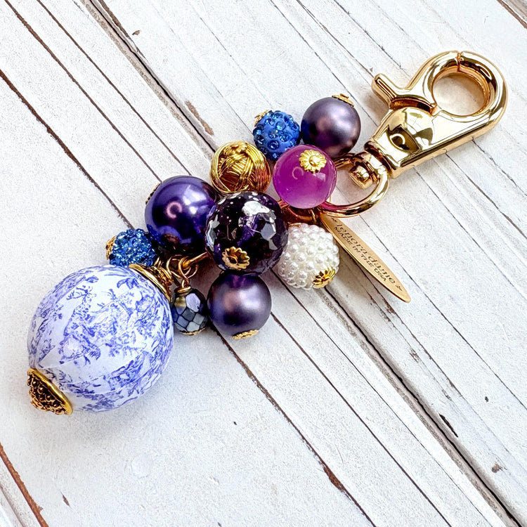 Beaded bag charm with a mix of bead styles in shades of purple, blue, gold and pearl. One larger round bead in a purple and white toile print hangs at the bottom and a gold trigger clasp on top of all of the beads. Brass oval hang tag near the trigger clasp that says "Lenora Dame Handmade in USA". Lenora Dame bag charm.