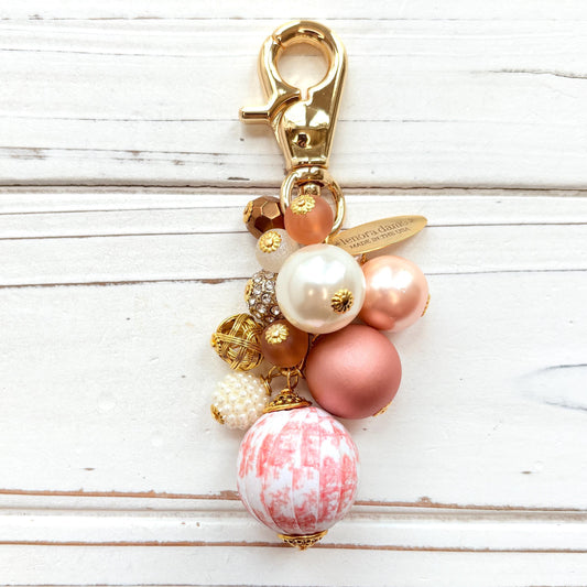 Beaded bag charm with a mix of bead styles in shades of pink, peach, gold and pearl. One larger round bead in a pink and white toile print hangs at the bottom and a gold trigger clasp on top of all of the beads. Brass oval hang tag near the trigger clasp that says "Lenora Dame Handmade in USA". Lenora Dame bag charm.