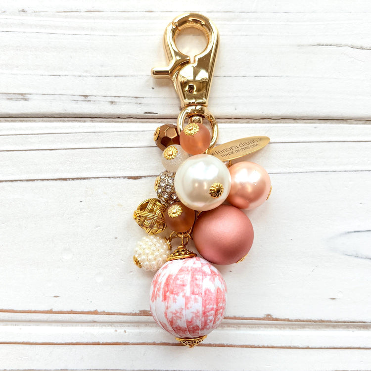 Beaded bag charm with a mix of bead styles in shades of pink, peach, gold and pearl. One larger round bead in a pink and white toile print hangs at the bottom and a gold trigger clasp on top of all of the beads. Brass oval hang tag near the trigger clasp that says "Lenora Dame Handmade in USA". Lenora Dame bag charm.