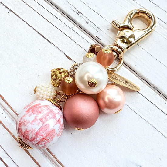 Beaded bag charm with a mix of bead styles in shades of pink, peach, gold and pearl. One larger round bead in a pink and white toile print hangs at the bottom and a gold trigger clasp on top of all of the beads. Brass oval hang tag near the trigger clasp that says "Lenora Dame Handmade in USA". Lenora Dame bag charm.