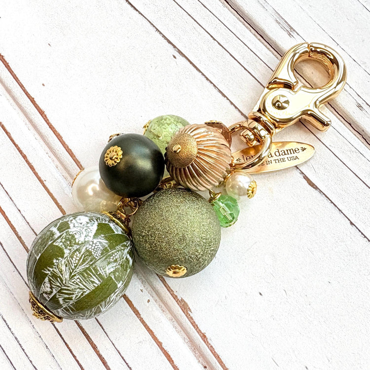 Beaded bag charm with a mix of bead styles in shades of green, gold, and pearl. One larger round bead in a army green and white toile print hangs at the bottom and a gold trigger clasp on top of all of the beads. Brass oval hang tag near the trigger clasp that says "Lenora Dame Handmade in USA". Lenora Dame bag charm.