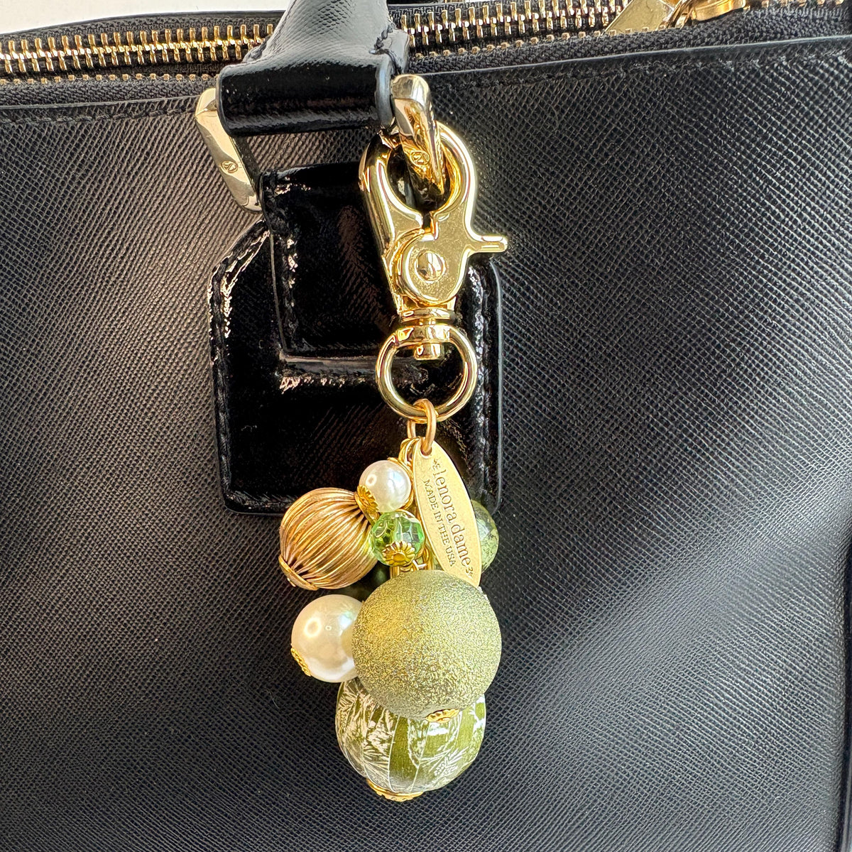 Beaded bag charm with a mix of bead styles in shades of green, gold, and pearl. One larger round bead in a army green and white toile print hangs at the bottom and a gold trigger clasp on top of all of the beads. Brass oval hang tag near the trigger clasp that says "Lenora Dame Handmade in USA". Lenora Dame bag charm.