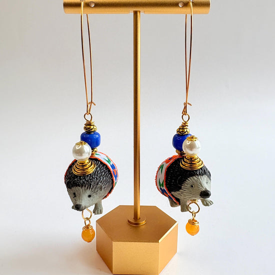 Cute hedgehog earrings with miniature black and gray hedgehog figurines wrapped in blue and orange floral ribbon trim hanging from long gold oval ear wires. Hedgehogs have gold cone and pearl hats and blue beads on their back. Lenora Dame earrings.