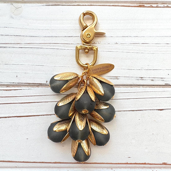 Beaded bag charm featuring matte black glass beads encased in gold plated brass floral leaf shaped bead caps. Gold trigger clasp and an oval hang tag that says Lenora Dame Made in USA. Lenora Dame bag charm.