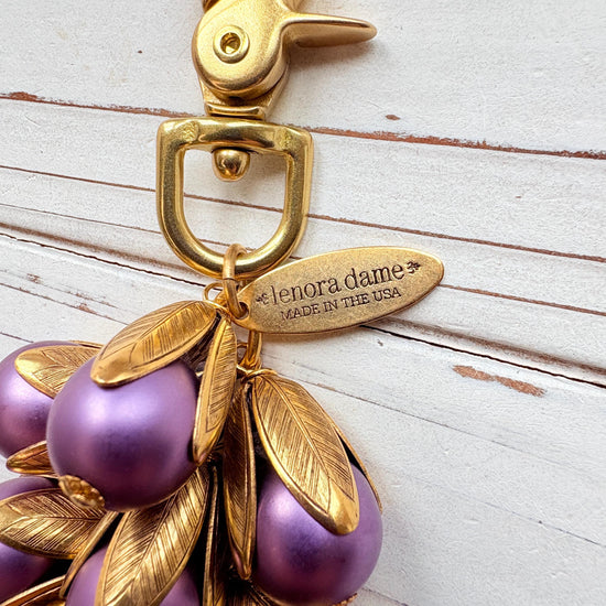 Gold bag charm with a trigger clasp at the top holding a cluster of lavender matte purple pearls encased in gold leaf bead caps. A gold oval tag imprinted with Lenora Dame Handmade in USA hangs at the top by the trigger clasp. Lenora Dame bag charms. Purple purse charm.