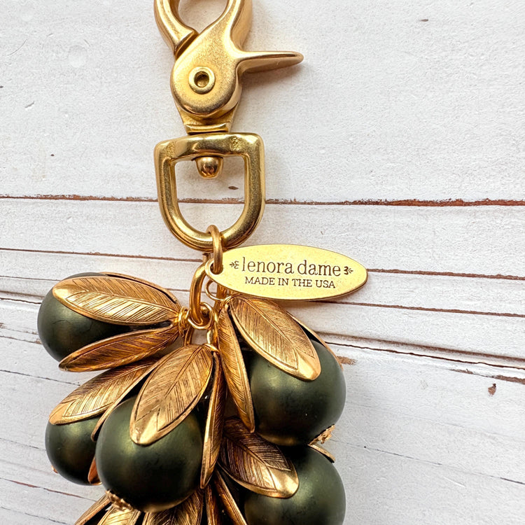 Gold bag charm with a trigger clasp at the top holding a cluster of matte hunter green pearls encased in gold leaf bead caps. A gold oval tag imprinted with Lenora Dame Handmade in USA hangs at the top by the trigger clasp. Lenora Dame bag charms. Green beaded purse charm.
