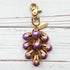 Gold bag charm with a trigger clasp at the top holding a cluster of lavender matte purple pearls encased in gold leaf bead caps. A gold oval tag imprinted with Lenora Dame Handmade in USA hangs at the top by the trigger clasp. Lenora Dame bag charms. Purple purse charm.