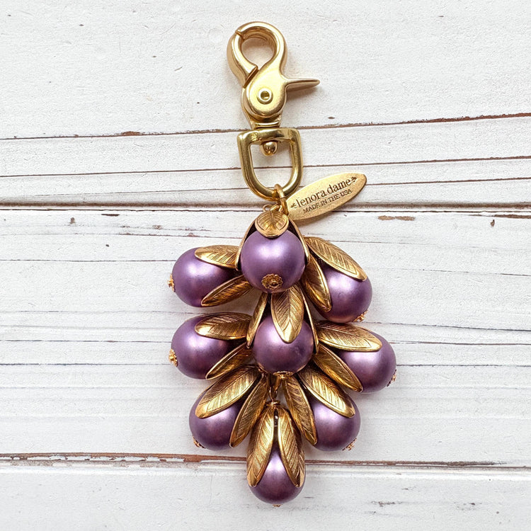 Gold bag charm with a trigger clasp at the top holding a cluster of lavender matte purple pearls encased in gold leaf bead caps. A gold oval tag imprinted with Lenora Dame Handmade in USA hangs at the top by the trigger clasp. Lenora Dame bag charms. Purple purse charm.