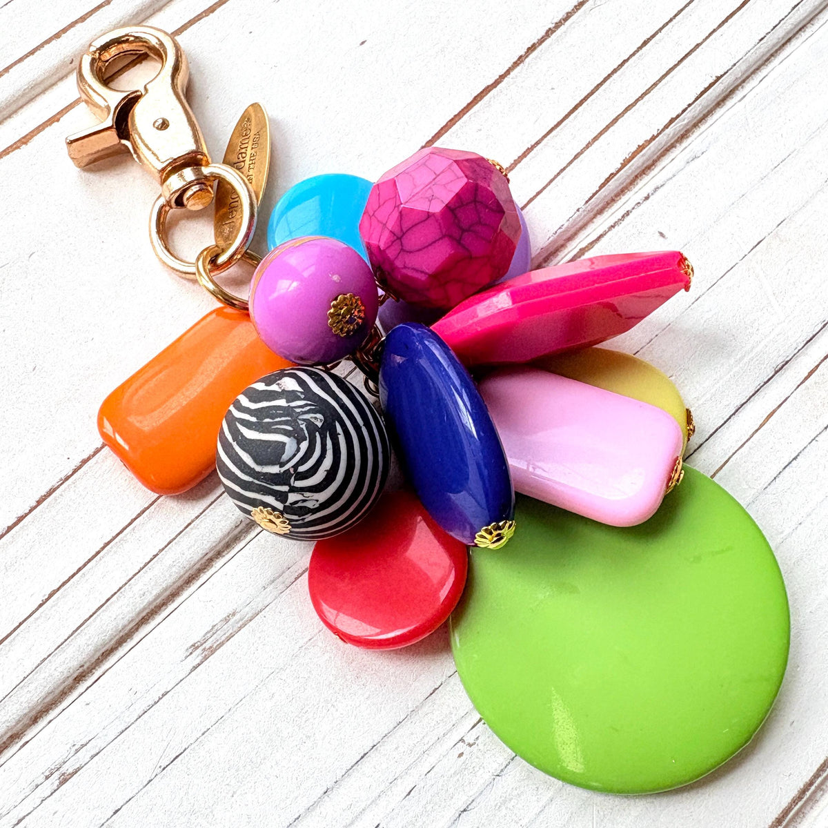 Colorful beaded bag charm with a mix of different shaped beads in a cluster under a gold trigger clasp with a gold oval charm that says Lenora Dame Made in USA. One round black and white zebra bead is among the beads. Lenora Dame bag charm.