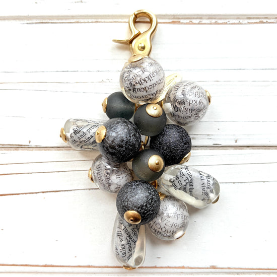 Black and white chunky bag charm with irregular shaped Lucite clear beads with a newspaper print inside hang with round black foil beads and black glass beads in a cluster under a gold trigger clasp. Lenora Dame bag charm.