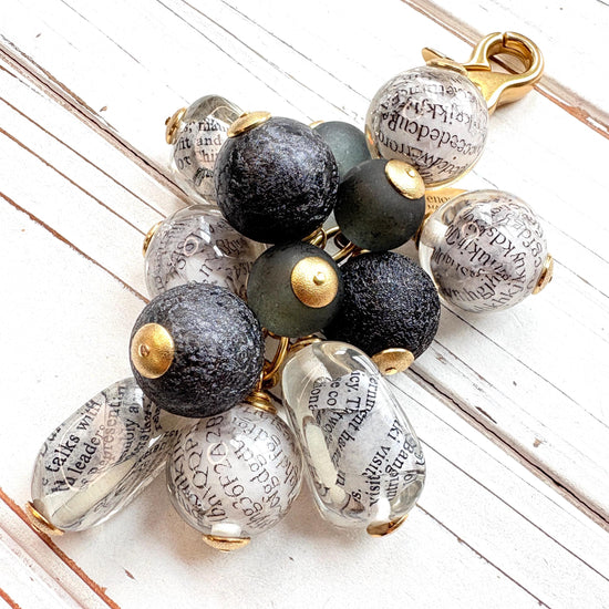 Black and white chunky bag charm with irregular shaped Lucite clear beads with a newspaper print inside hang with round black foil beads and black glass beads in a cluster under a gold trigger clasp. Lenora Dame bag charm.